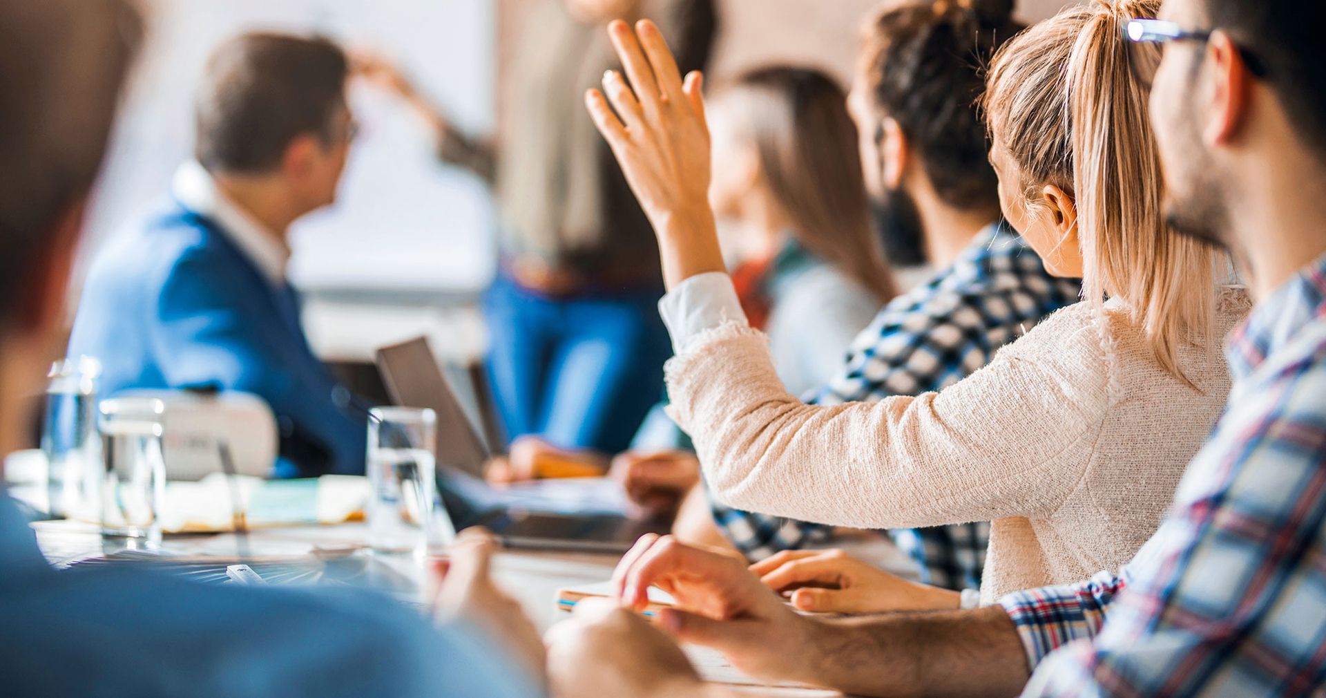 Mehrere Personen sitzen bei einem Seminar an einem Tisch und eine Frau hebt ihre Hand