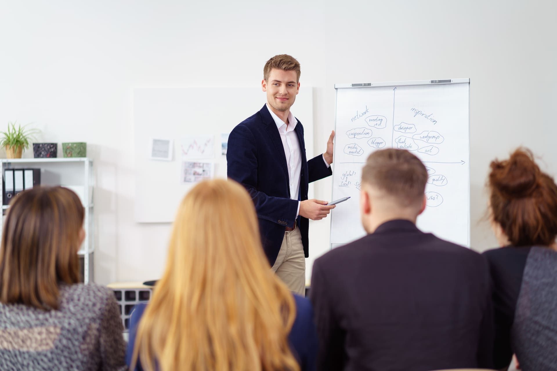 Ein Mann im Hintergrund hält einen Vortrag und steht dabei neben einem Flipchart